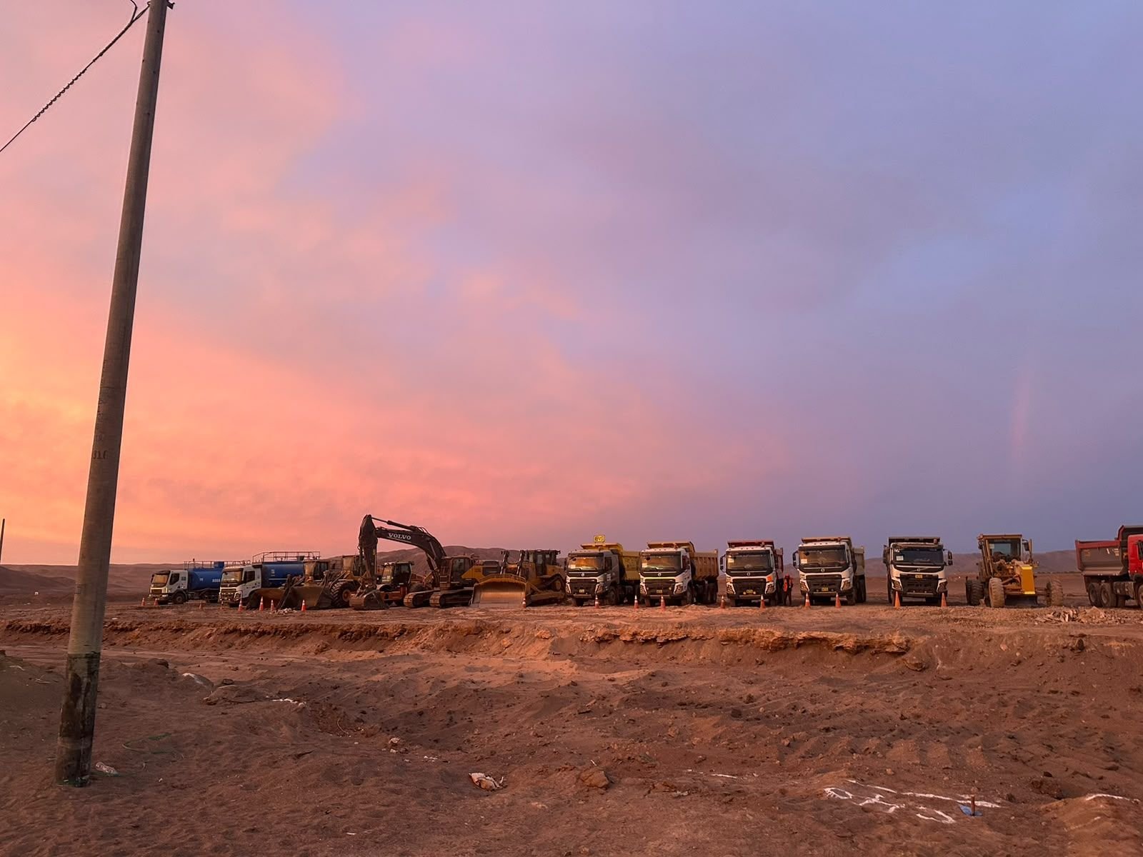 Heavy equipment and dump trucks lined up at sunset on desert terrain.